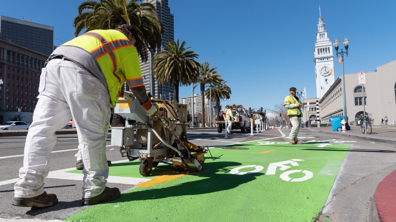 Person in neon yellow shirt using machinery to paint bike lane depictions on road asphalt, Ferry Building and palm trees in background 