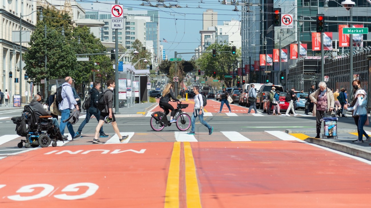 Red transit lane with pedestrians and wheelchair user in crosswalk, cyclist riding adjacent to pedestrians