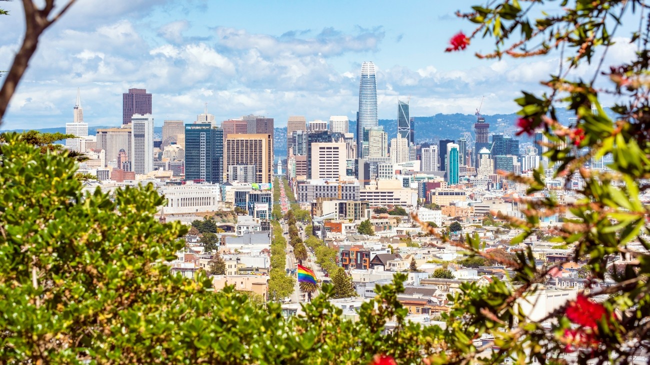 A view of Downtown San Francisco, looking down Market Street, seen from up on a hill and through bushes and trees. Large, fluffy, white clouds populate the blue sky