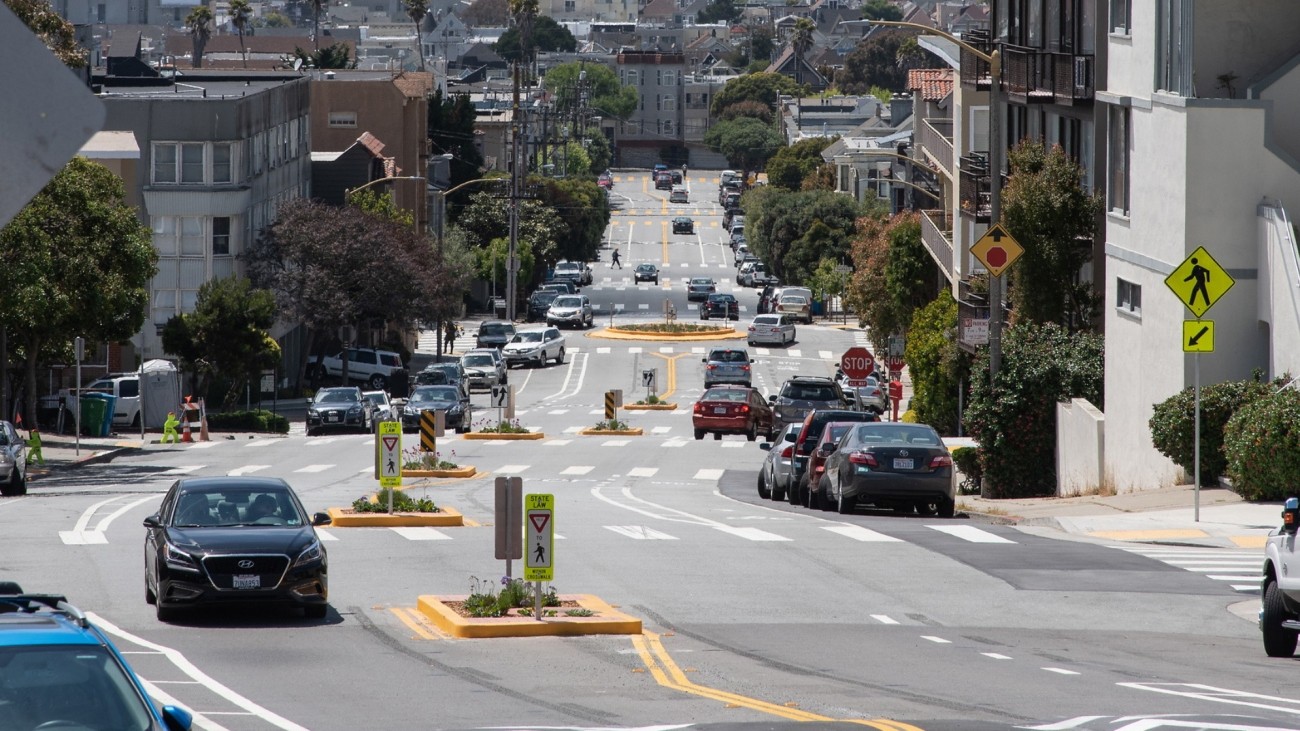 Street with crosswalks, landscaped medians, speed humps, and a bike lane. Various signs along the street indicate to yield to pedestrians. There are cars driving on the street, and cars are parked along its sides.