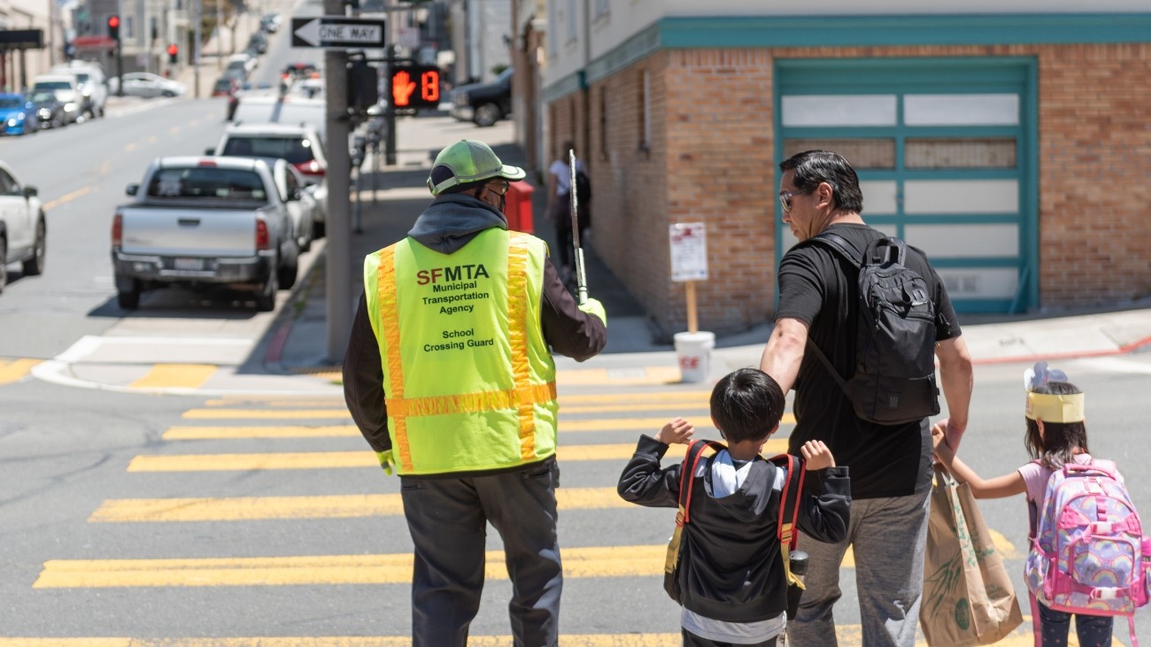 Crossing guard in neon yellow vest holds up stop sign at a crosswalk, man with two children is beside crossing guard and crossing the street