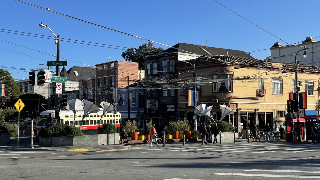 Street-level view of sunny Jane Warner Plaza and intersection of Market and Castro; a streetcar is turning from 17th onto Market Street.