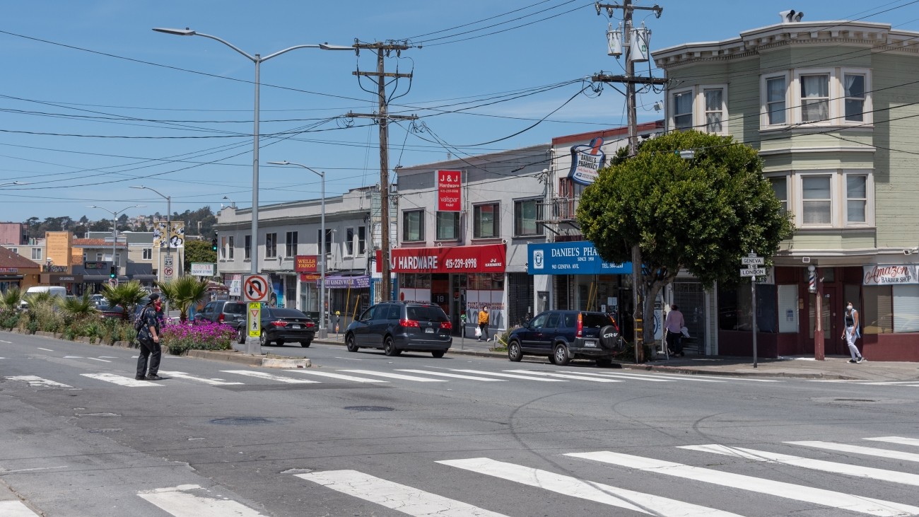 Intersection near at Geneva Ave and London Street, off of Mission Street. Pedestrian is crossing is on marked crosswalk and other pedestrians are on the sidewalk, cars are parked.