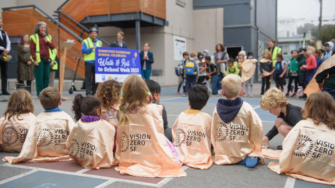 Children sitting on playground floor wearing gold toned capes that read "Vision Zero SF" with the Vision Zero SF logo, adults and other children are standing and blurred in the background