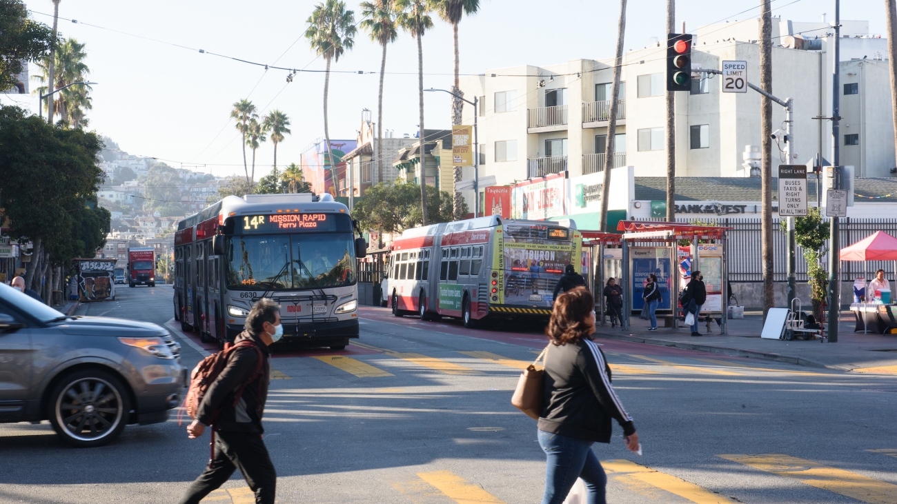 Pedestrians walk across Mission Street, 14R Muni line is driving up, and palm trees line the street in the background