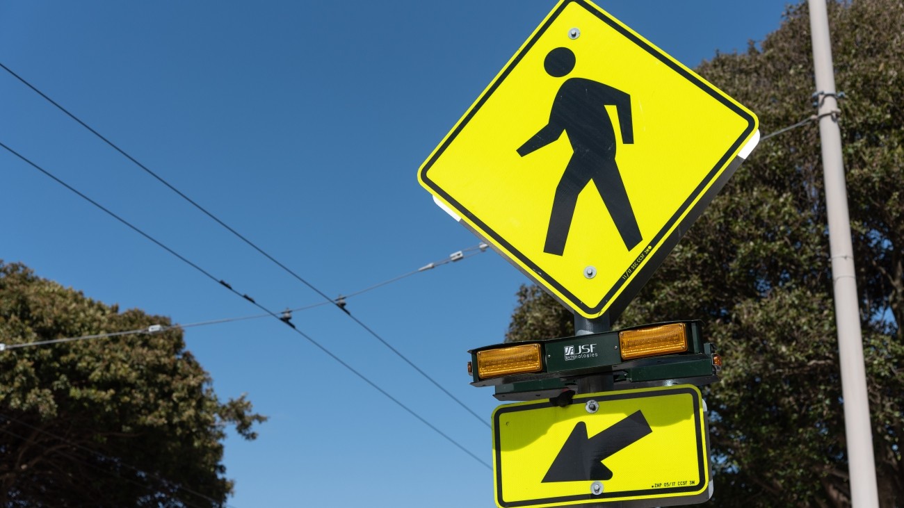 Diamond shaped yellow sign with pedestrian symbol, rectangular lights below it which are intended to flash to when pedestrians cross to increase pedestrian visibility 