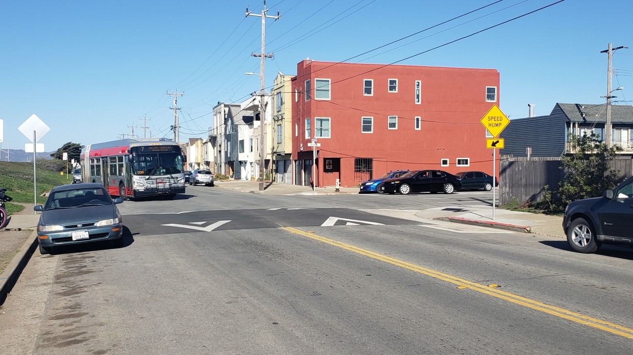 The intersection of Great Highway and Ortega on a sunny day. The 7 Muni bus approaches the intersection, which sports a new speed table crossing.