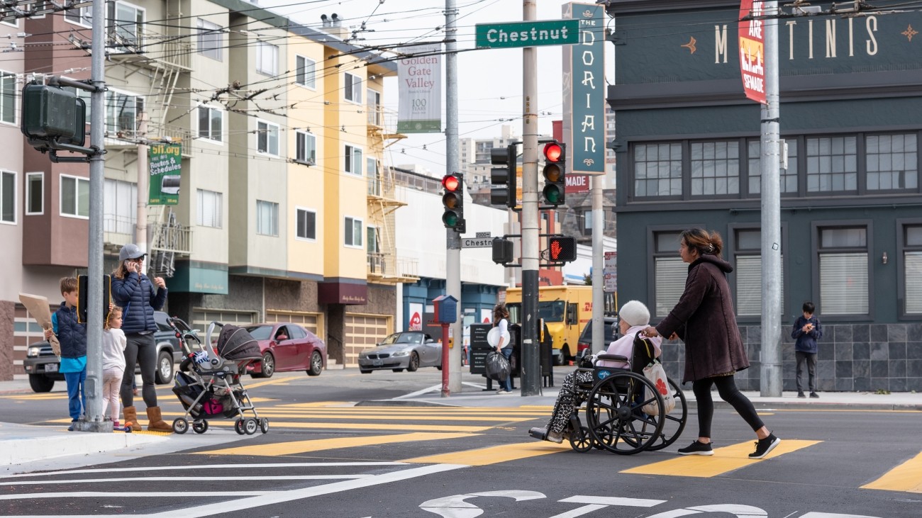 A parent with two kids and a stroller wait at the corner of Chestnut and Fillmore, while a young woman pushes an elderly woman in a wheelchair in the crosswalk.