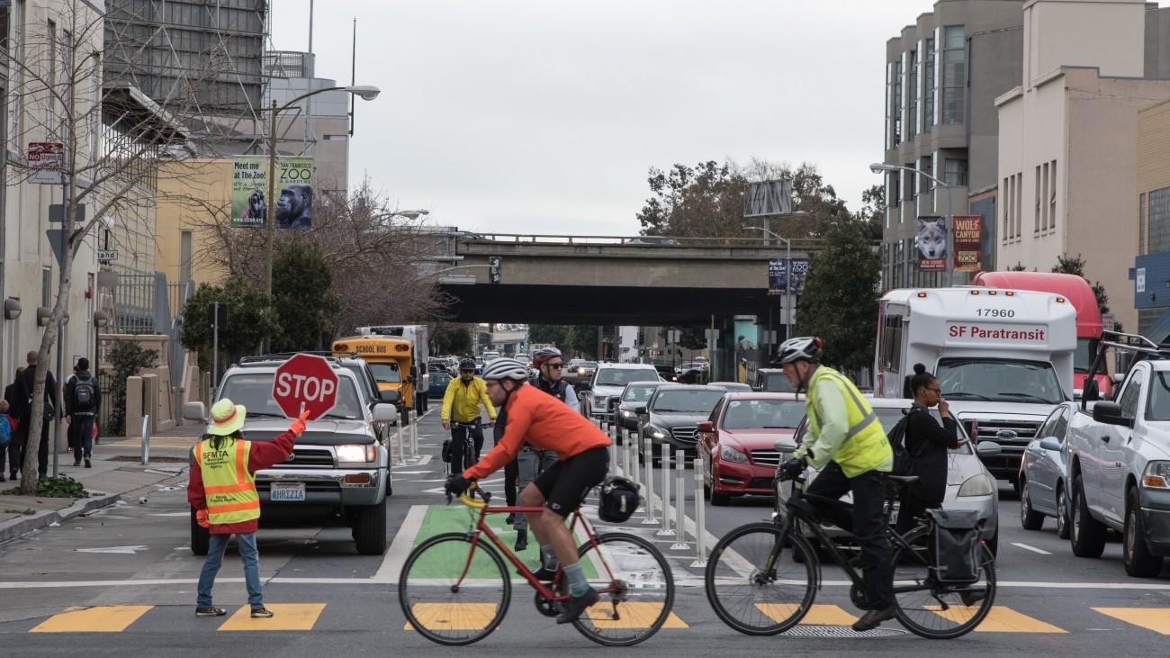 Cyclists and pedestrians crossing with cars stopped and a crossing guard in the crosswalk. 