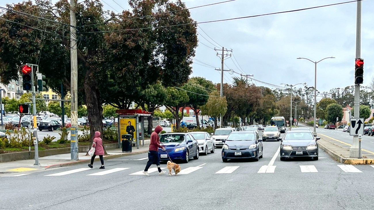 People cross Brotherhood Way in a crosswalk near a bus shelter while car traffic waits. A Muni bus can be seen in the background.