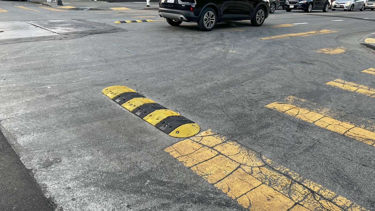 Eye-level view of an intersection, the pavement streaked with sideshow tire markings. Black and yellow striped sideshow deterrence humps, about 4 feet long, 1 foot wide, and a few inches high, protrude into the intersection from the corners and between the two lanes of each street.