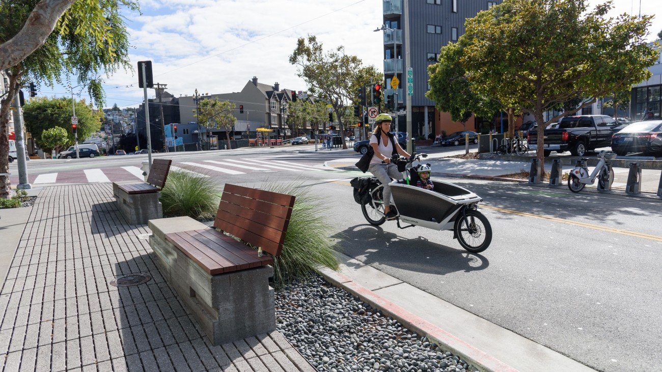 Seating and planters with adult and child in cargo bike riding by on the road