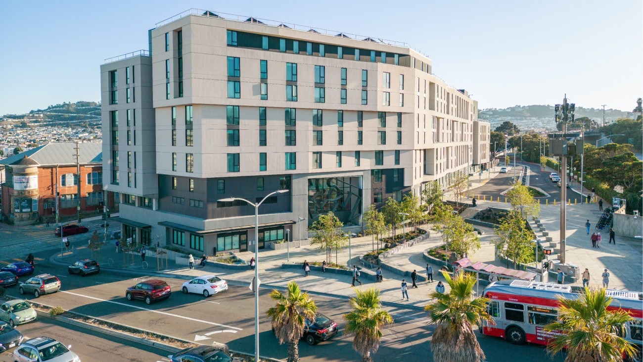 The plaza and affordable housing development at Balboa Park Upper Yard, seen from above and across Geneva Avenue. There are people walking in the plaza and a Muni bus in the street.