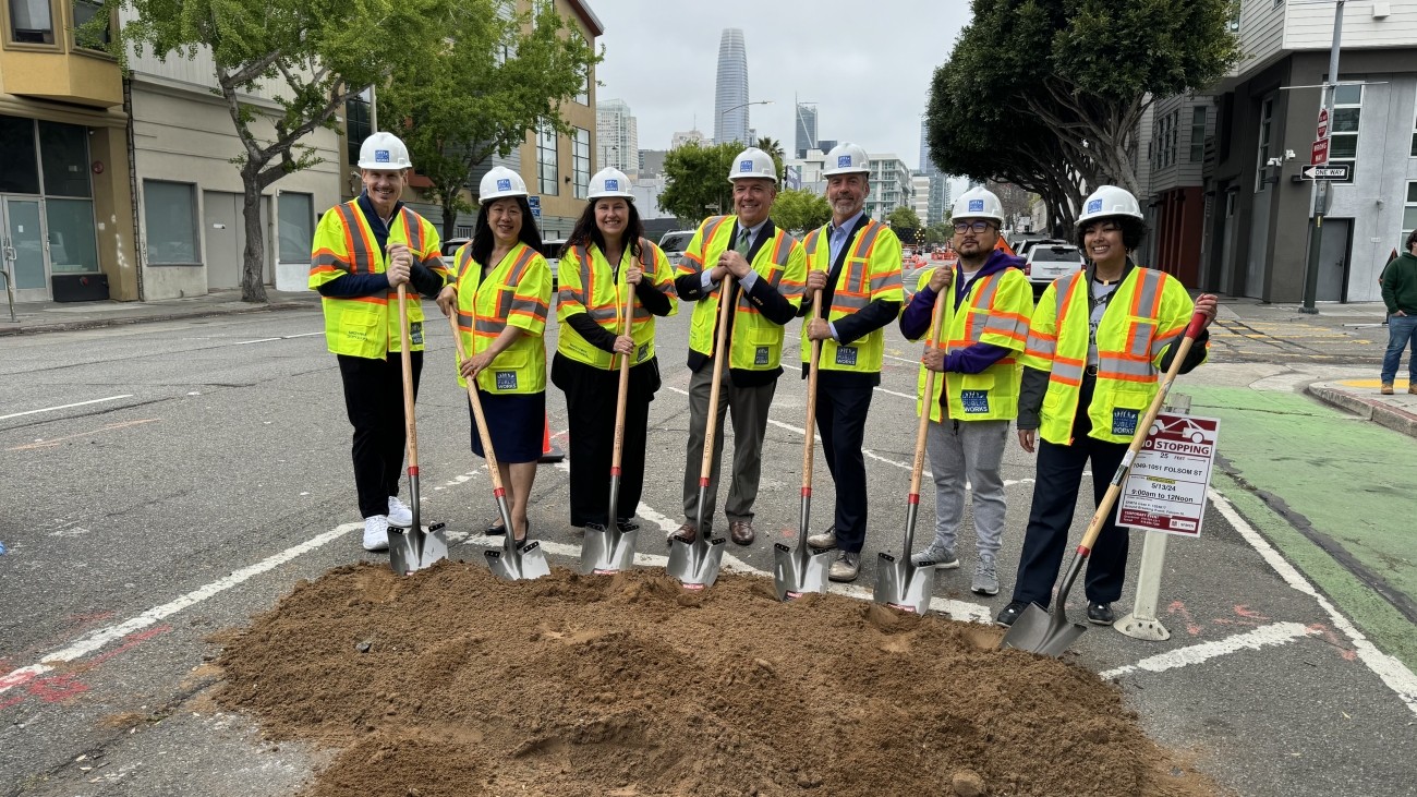 People are standing in the roadway wearing hard hats and safety vests. They are holding shovels in front a dirt pile. Trees and building are seen in the background.