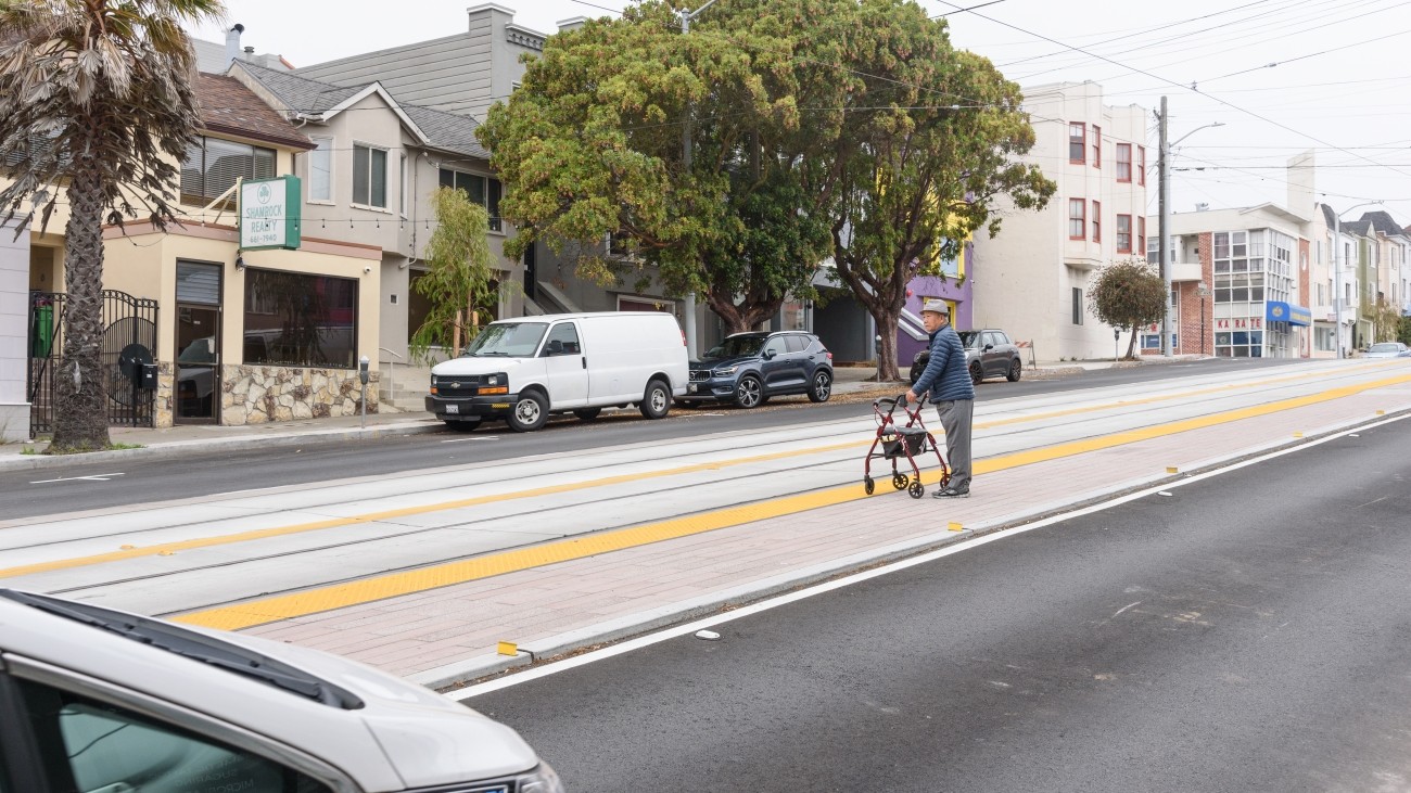 A senior citizen wearing a hat holding a walker waiting for L Taraval at new boarding platform on Taraval Street