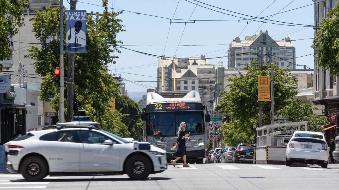 A Waymo vehicle driving in an intersection at the foreground, with a pedestrian and Muni bus in the background