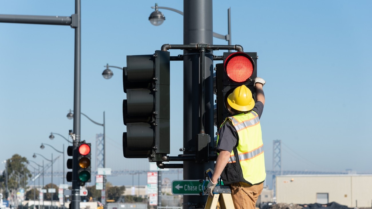 A person in a yellow vest and construction helmet on a ladder to reach a traffic signal