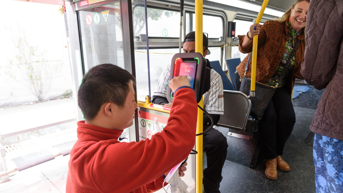 A person wearing an orange jacket boarding a Muni bus and tapping their Clipper card on the reader