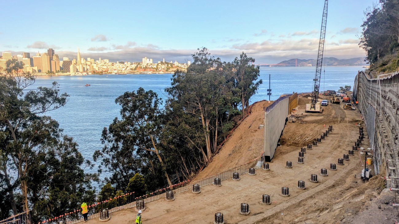 Workers use heavy machinery to grade the earth and pour concrete footings on Yerba Buena Island. The San Francisco skyline is visible in the background