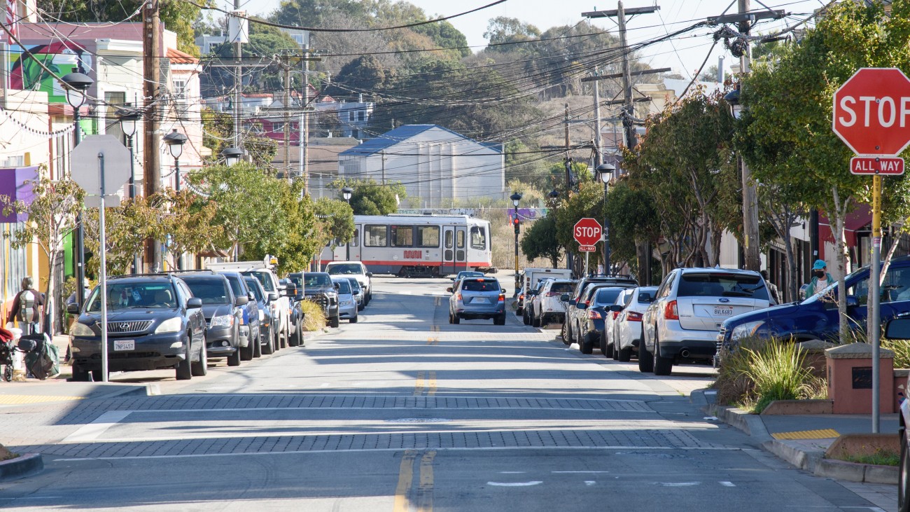 View of a road in Visitacion Valley, with a train in the distance and cars parked along the side