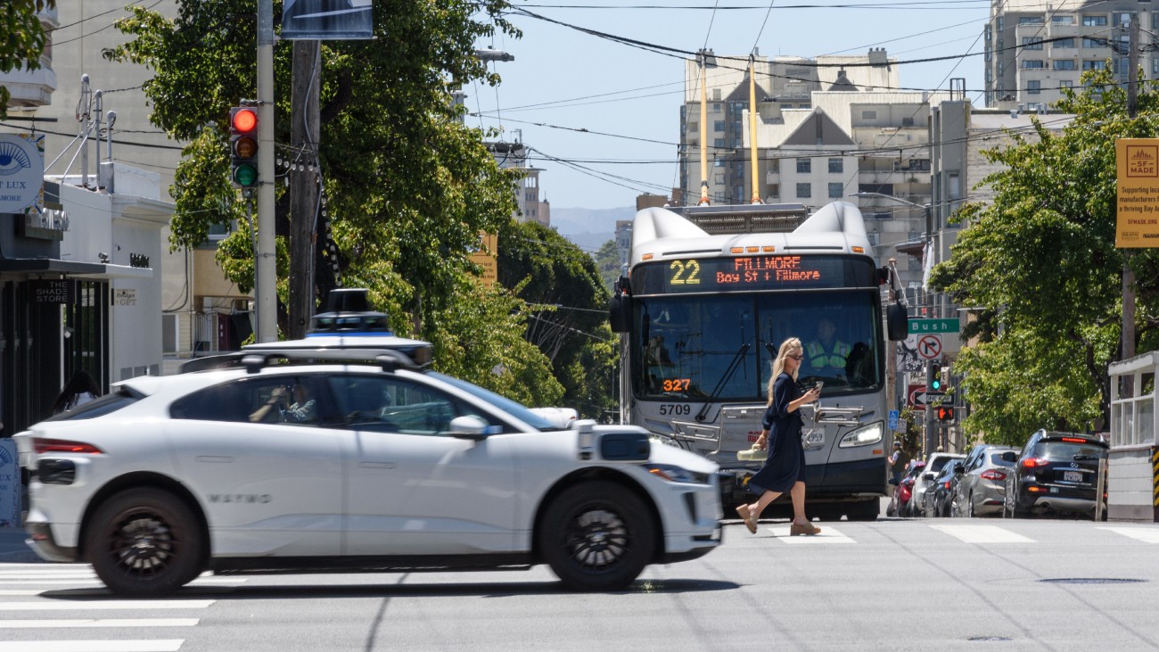 A Waymo autonomous vehicle drives through an intersection, while a 22 Fillmore Muni bus waits in the background.