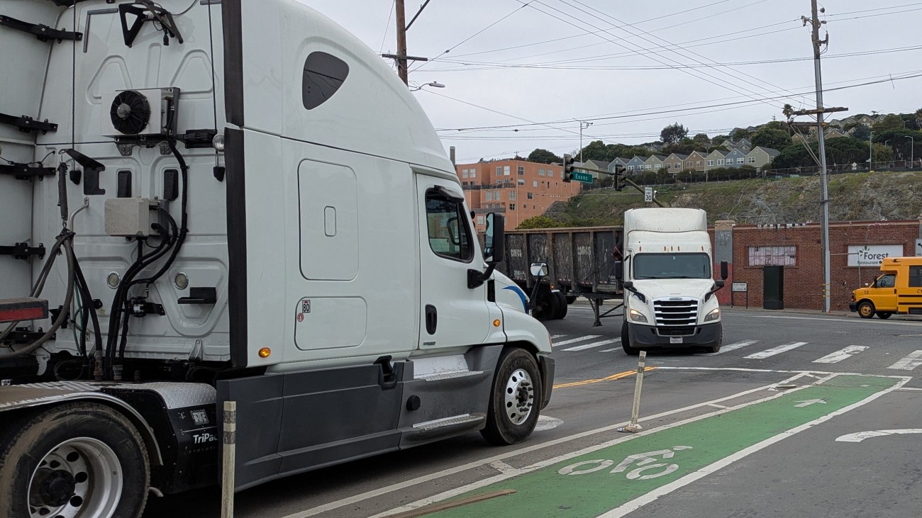 Two large trucks pass each other on Evans Avenue and Cesar Chavez St. A green-painted bike lane is in the foreground.