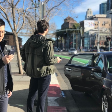 People standing on a sidewalk with phones and a ride-hail vehicle dropping someone off