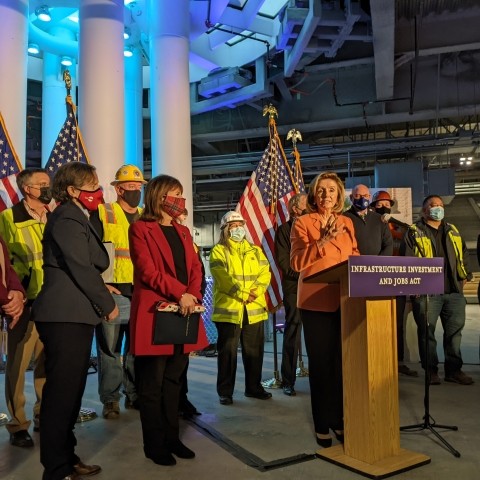 Speaker Pelosi speaking at the podium with city officials and agency staff gathered around.