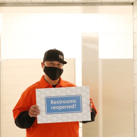District Works worker holds sign saying "Restrooms Reopened!" in front of the Powell BART Station restroom.