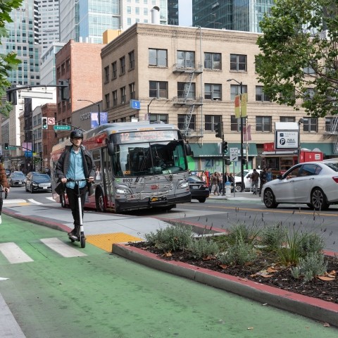 people on bike and scooter on green lane, transit boarding island with landscaping, and Muni bus