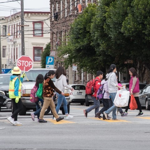 Children and adults crossing the street with a crossing guard along Broadway Street
