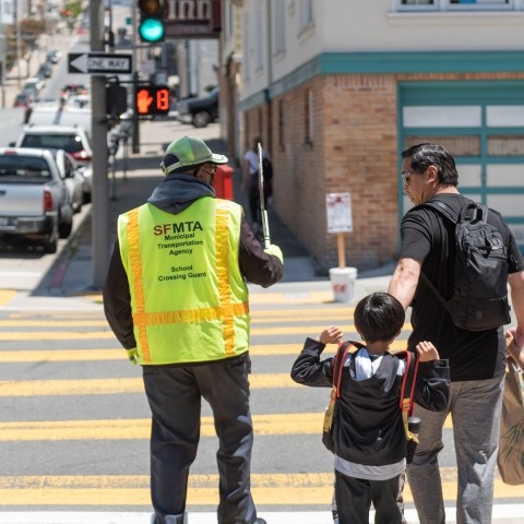 Crossing guard in neon yellow vest holds up stop sign at a crosswalk, man with two children is beside crossing guard and crossing the street