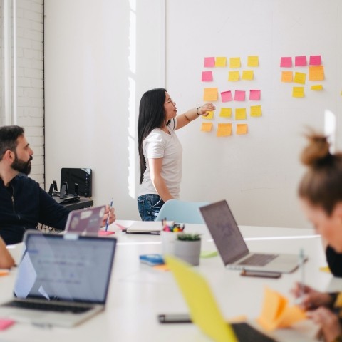 People sitting in front of laptops around a table, person standing at wall and pointing to post it notes