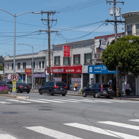 Intersection near at Geneva Ave and London Street, off of Mission Street. Pedestrian is crossing is on marked crosswalk and other pedestrians are on the sidewalk, cars are parked. 