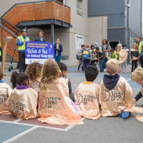 Children sitting on playground floor wearing gold toned capes that read "Vision Zero SF" with the Vision Zero SF logo, adults and other children are standing and blurred in the background