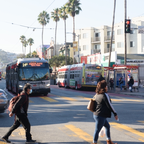 Pedestrians walk across Mission Street, 14R Muni line is driving up, and palm trees line the street in the background