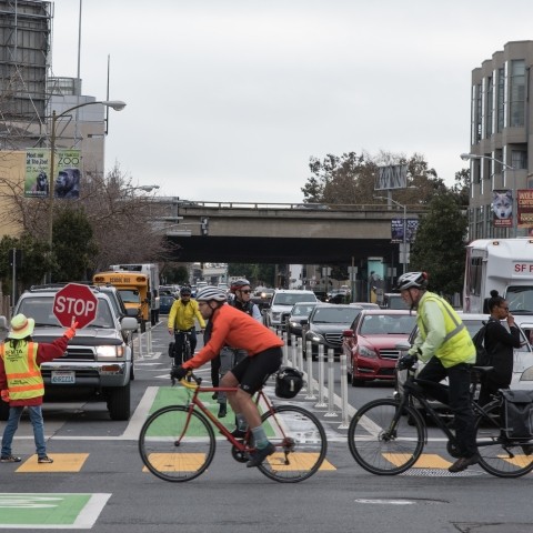 Cyclists and pedestrians crossing with cars stopped and a crossing guard in the crosswalk. 