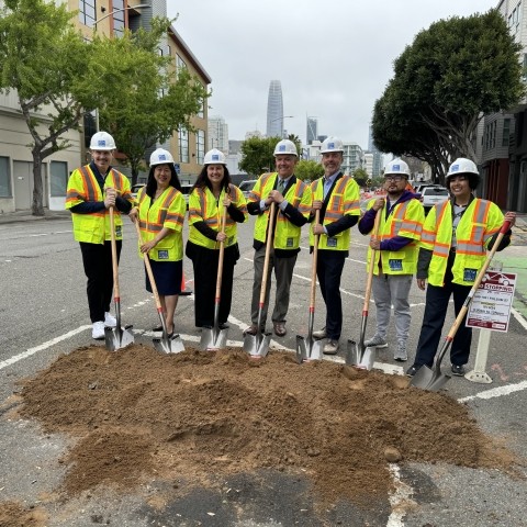 People are standing in the roadway wearing hard hats and safety vests. They are holding shovels in front a dirt pile. Trees and building are seen in the background. 