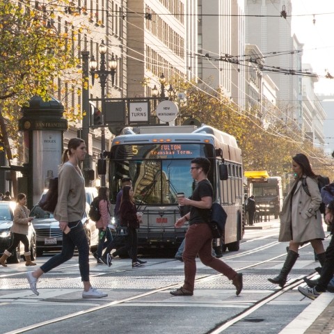 Pedestrians crossing the street with a Muni bus and vehicles waiting at the intersectionn