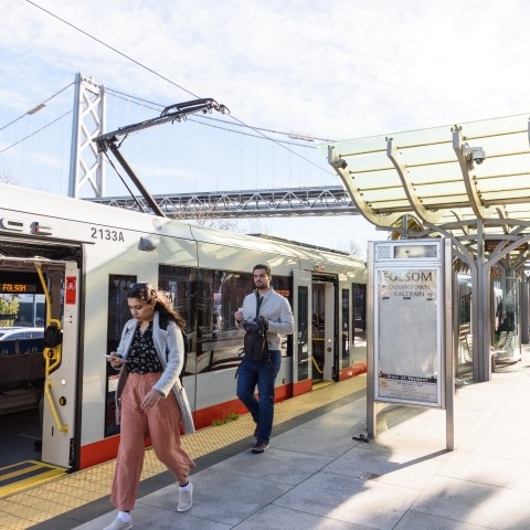 People offboarding a Muni metro train onto a boarding platform, with the Bay Bridge in the background