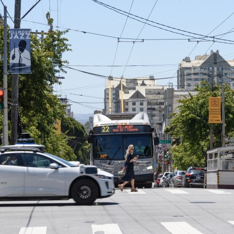 A Waymo vehicle driving in an intersection at the foreground, with a pedestrian and Muni bus in the background