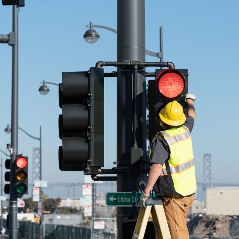 A person in a yellow vest and construction helmet on a ladder to reach a traffic signal