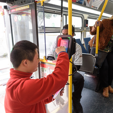 A person wearing an orange jacket boarding a Muni bus and tapping their Clipper card on the reader