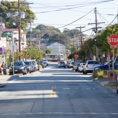 View of a road in Visitacion Valley, with a train in the distance and cars parked along the side