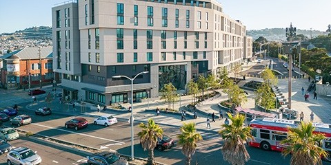 The plaza and affordable housing development at Balboa Park Upper Yard, seen from above and across Geneva Avenue. There are people walking in the plaza and a Muni bus in the street.