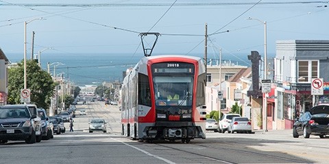 A new Muni light rail vehicle in operation on a sunny day with the ocean in the background.