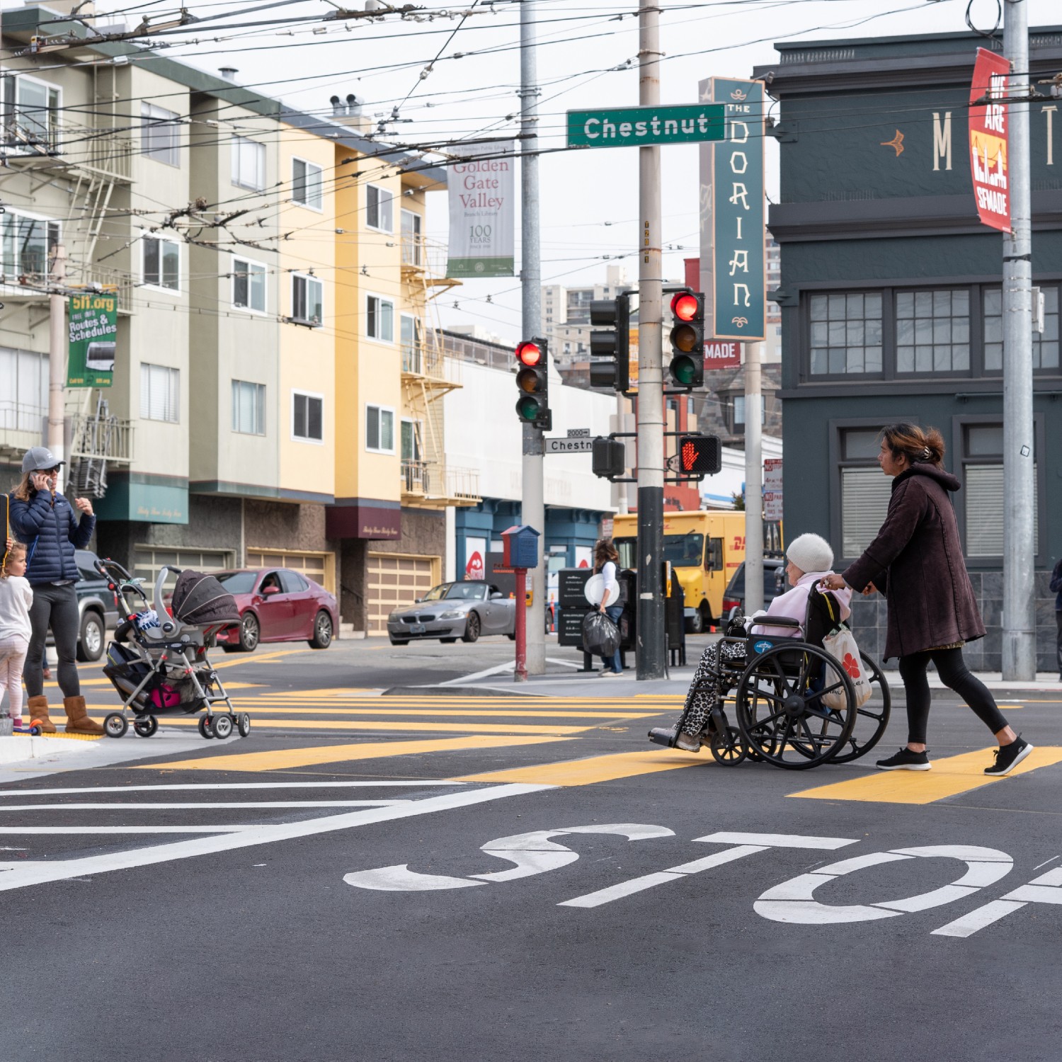 A parent with two kids and a stroller wait at the corner of Chestnut and Fillmore, while a young woman pushes an elderly woman in a wheelchair in the crosswalk.